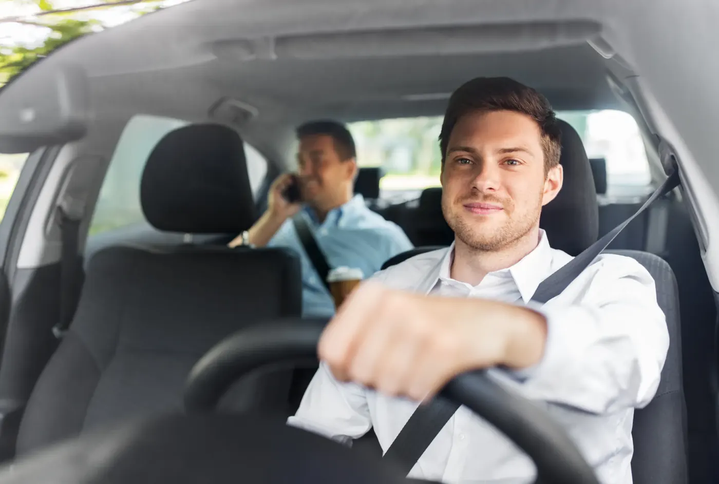 A driver in a white shirt wearing a fastened seat belt at the wheel of a car, with a second occupant belted in the rear seat behind him.