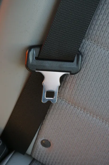 Metal seat belt tongue plate held in a black webbing guide against a light-coloured vehicle seat.