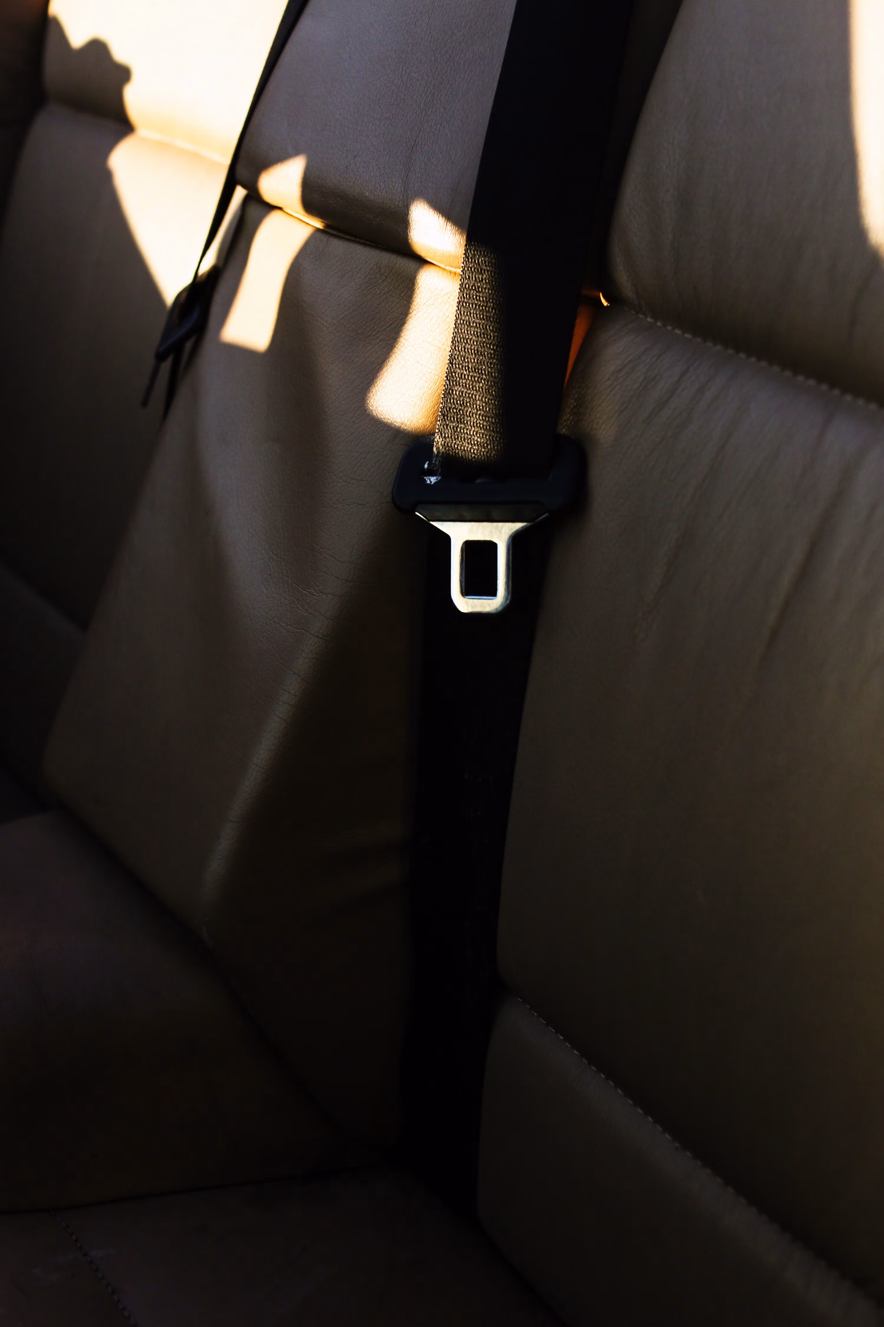 Rear bench seat in tan leather with a dark seat belt running vertically past the tongue plate, lit by a shaft of daylight.
